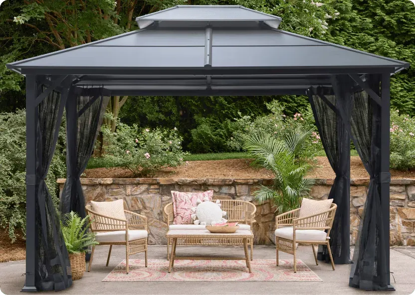 Dark aluminium hardtop gazebo with a two-tier vented roof and mosquito netting panels, over a wicker seating set on a stone patio