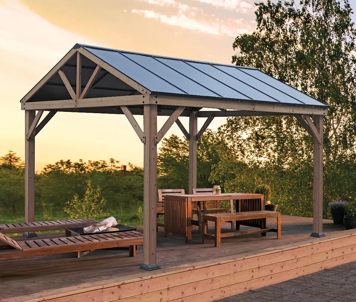 Wood-frame gazebo with a dark metal hardtop roof sheltering a deck dining table and benches at sunset