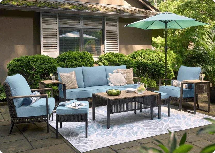Patio conversation set with a wicker sofa and two armchairs in light blue cushions, a slatted coffee table and a teal market umbrella on a stone patio