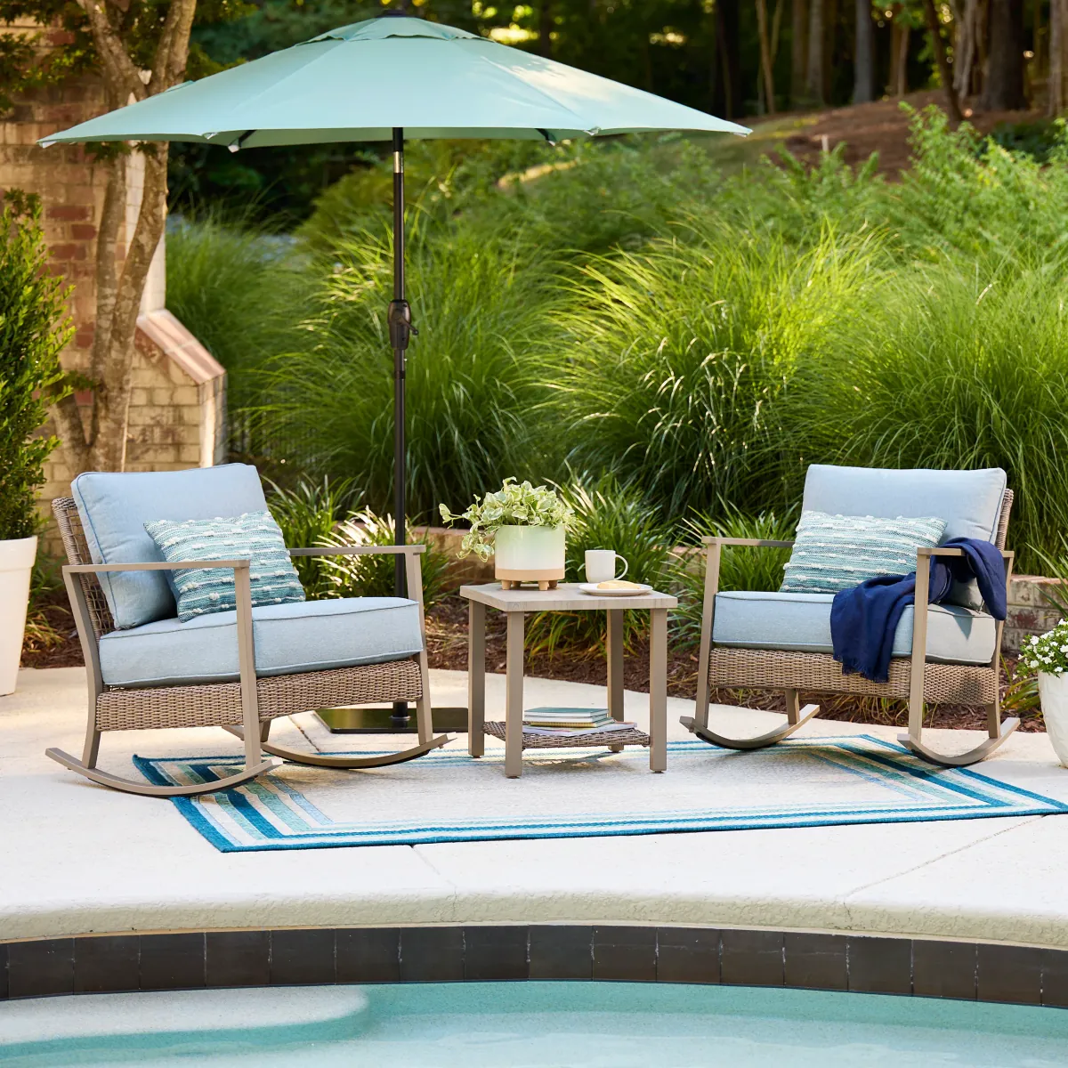 Two wicker patio rocking chairs with light blue cushions beside a side table and a teal market umbrella on a poolside patio