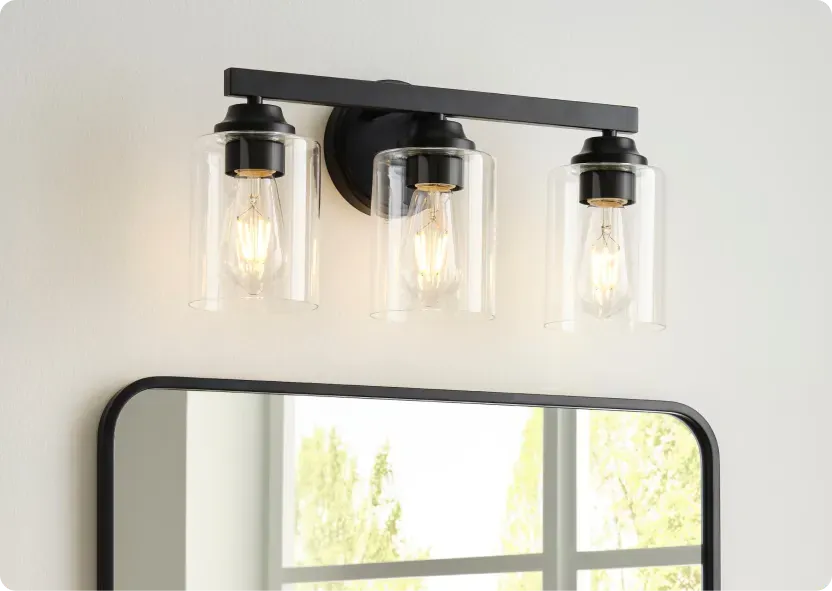 Black three-light vanity bar with clear glass shades and exposed filament bulbs, lit above a rounded black-framed bathroom mirror